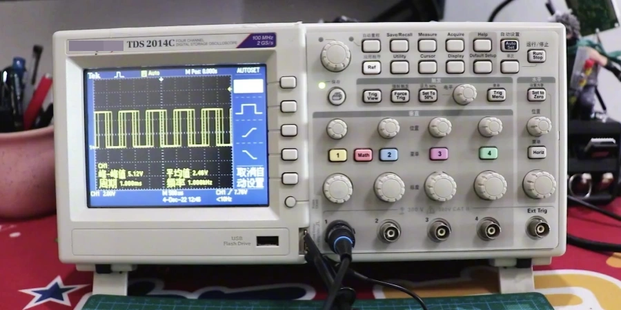 Digital oscilloscope on a workbench, screen showing yellow square wave/pulse signals in automatic mode, with grid background, measurement readouts (period, average voltage), and channel settings visible. Front panel with buttons, knobs, and connected probe on a cutting mat.