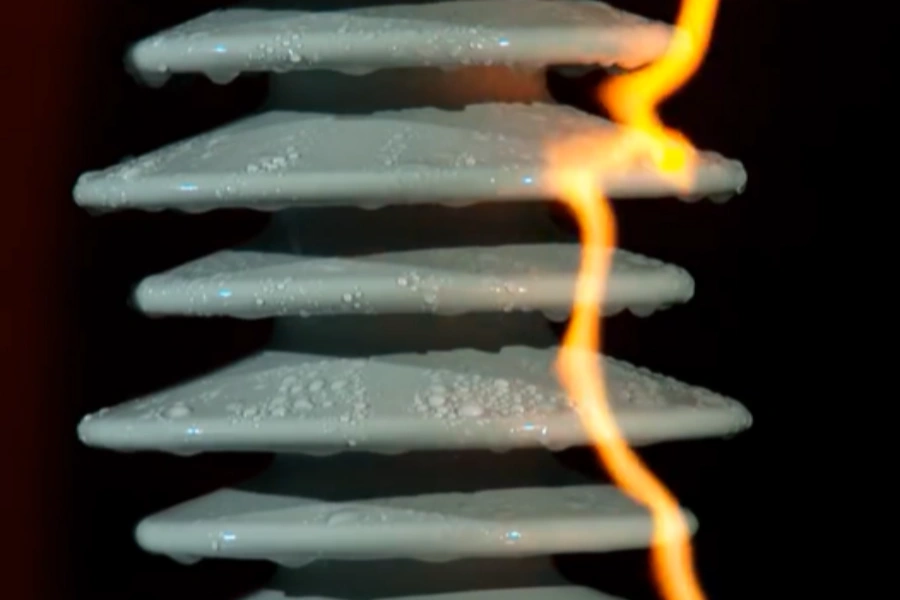 Close-up view of a white ceramic high-voltage insulator with multiple stacked discs covered in water droplets, showing surface tracking with a bright orange electrical arc flashing along the right side against a dark background